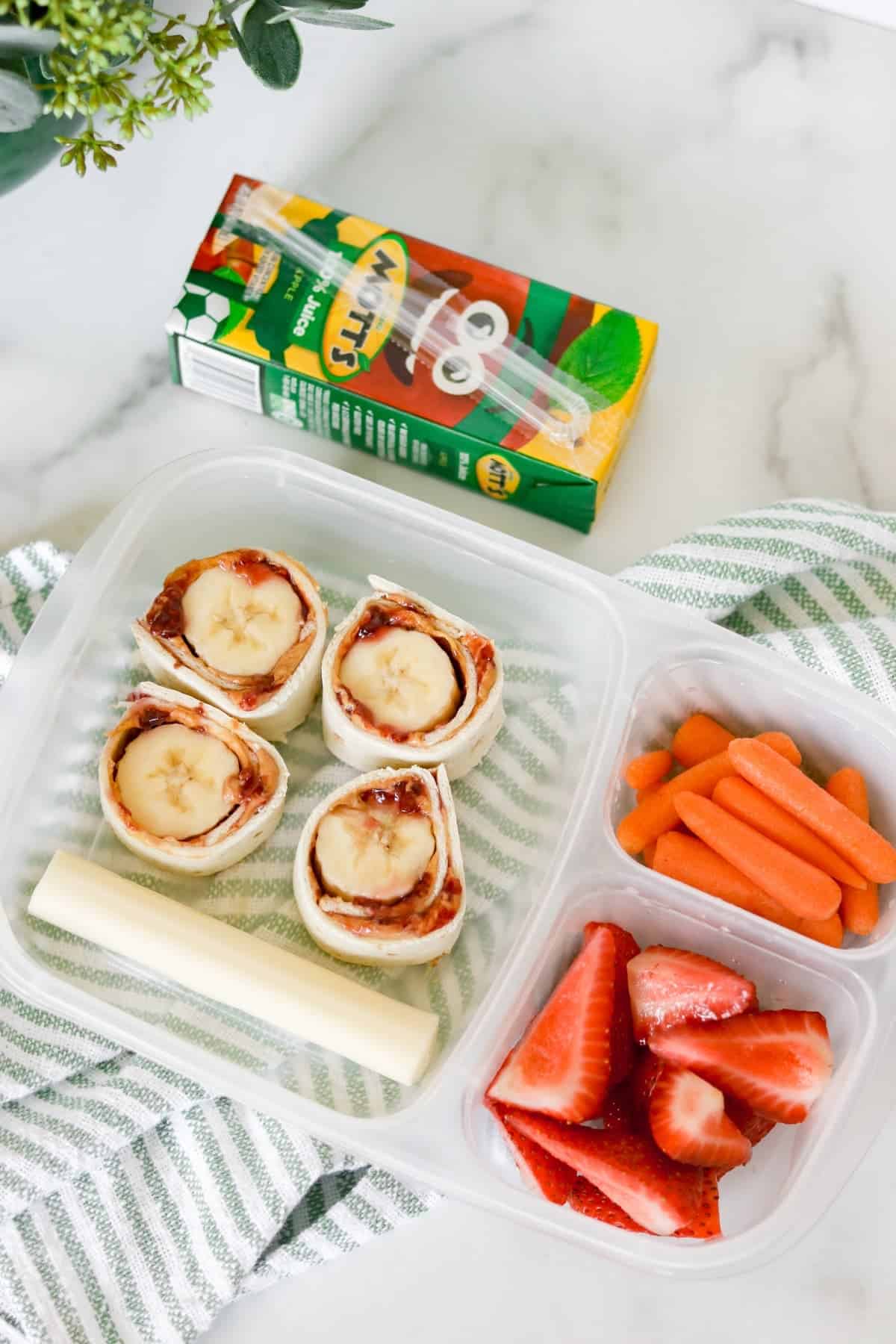 A kids lunch featuring banana and peanut butter roll-ups, baby carrots, sliced strawberries, a cheese stick, and a juice box on a striped cloth—perfect for pool snacks or quick pool lunch ideas.