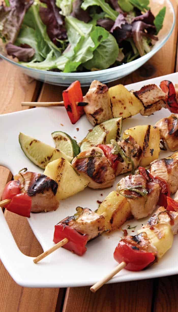 Grilled chicken, pineapple, and red pepper skewers on a white plate, perfect for summer grilling, with lime wedges and a mixed green salad in the background.