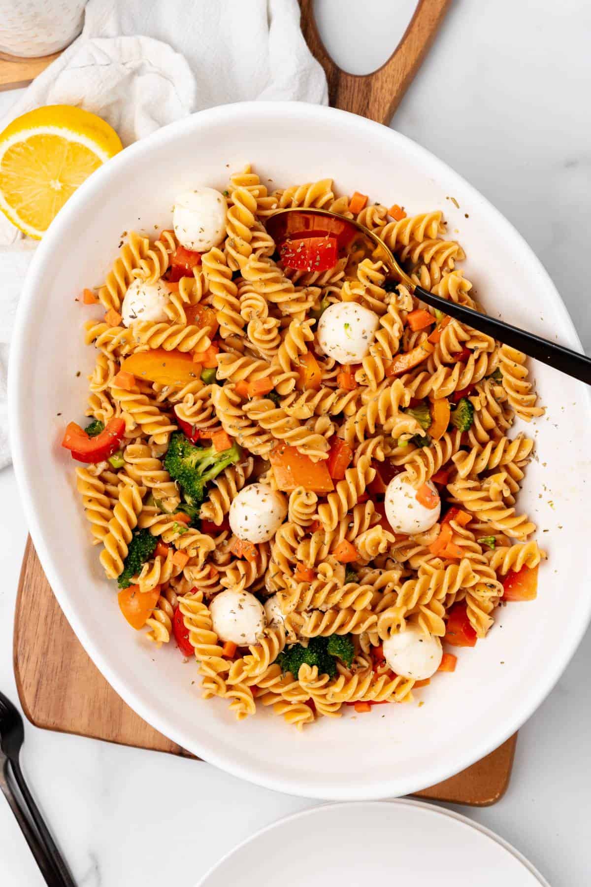 A white oval dish filled with fusilli pasta, cherry tomatoes, broccoli, sliced bell peppers, and mozzarella balls—perfect as a side dish for your graduation party, served with sleek black utensils on the side.