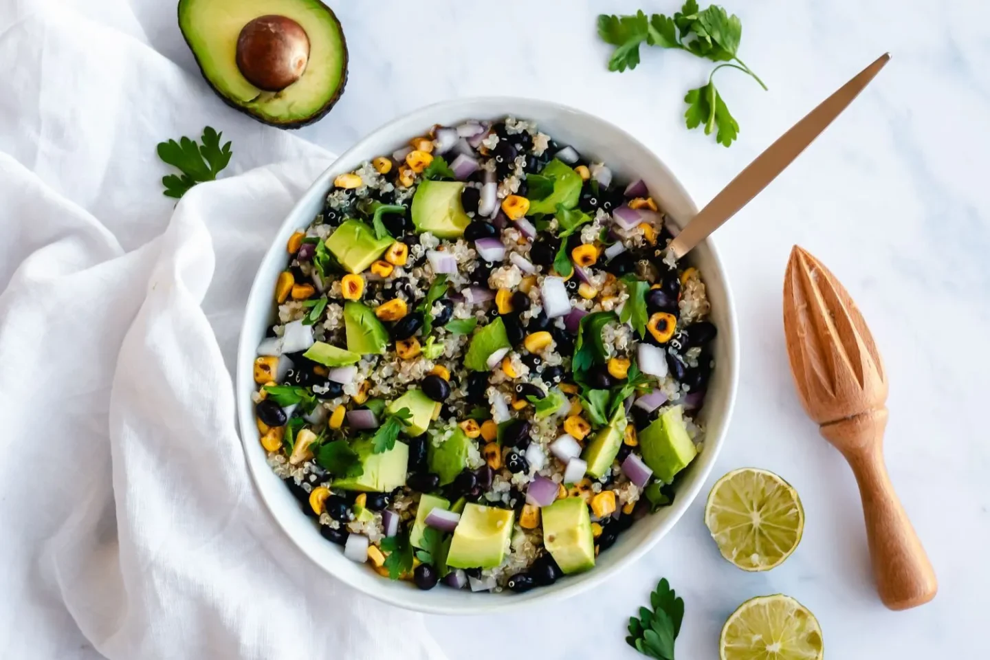 A bowl of quinoa salad with diced avocado, onion, black beans, corn, and herbs—perfect for cooler meals or as easy boat food—sits next to a halved avocado, parsley, lime halves, and a wooden citrus reamer on a white surface.