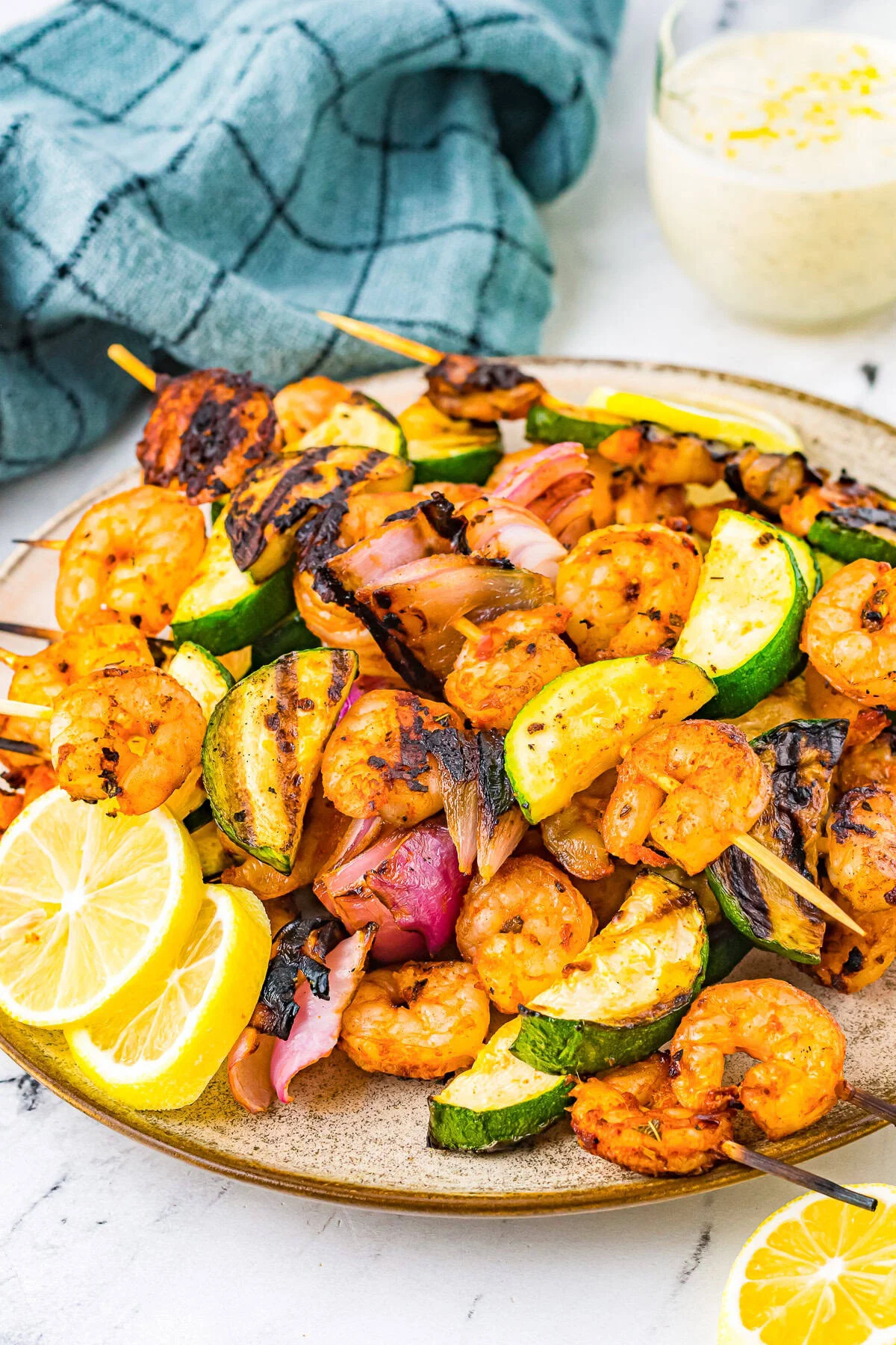 Grilled shrimp and vegetable skewers, featuring zucchini and red onion, arranged on a plate with lemon slices—perfect for a light summer dinner. A blue cloth and a bowl of sauce sit in the background.