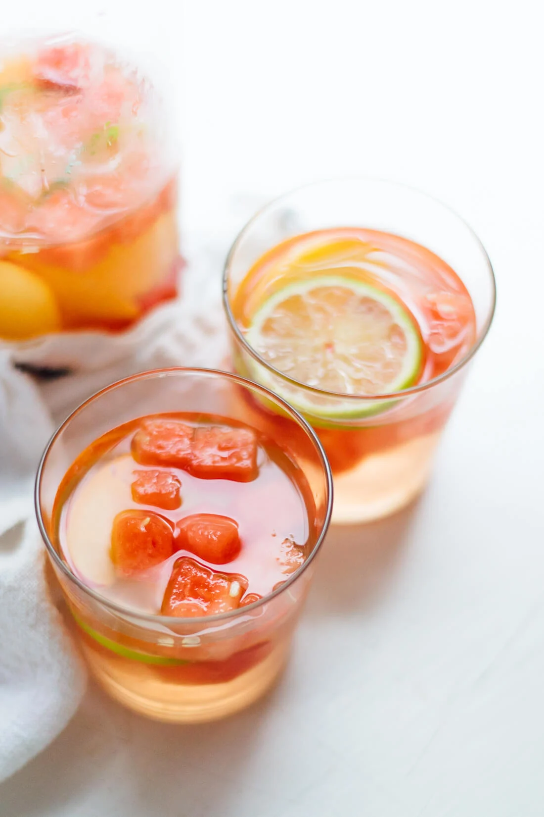 Two glasses of light orange summer sangria with ice cubes and lime slices sit on a white surface, while a pitcher of the refreshing beverage lingers in the background.