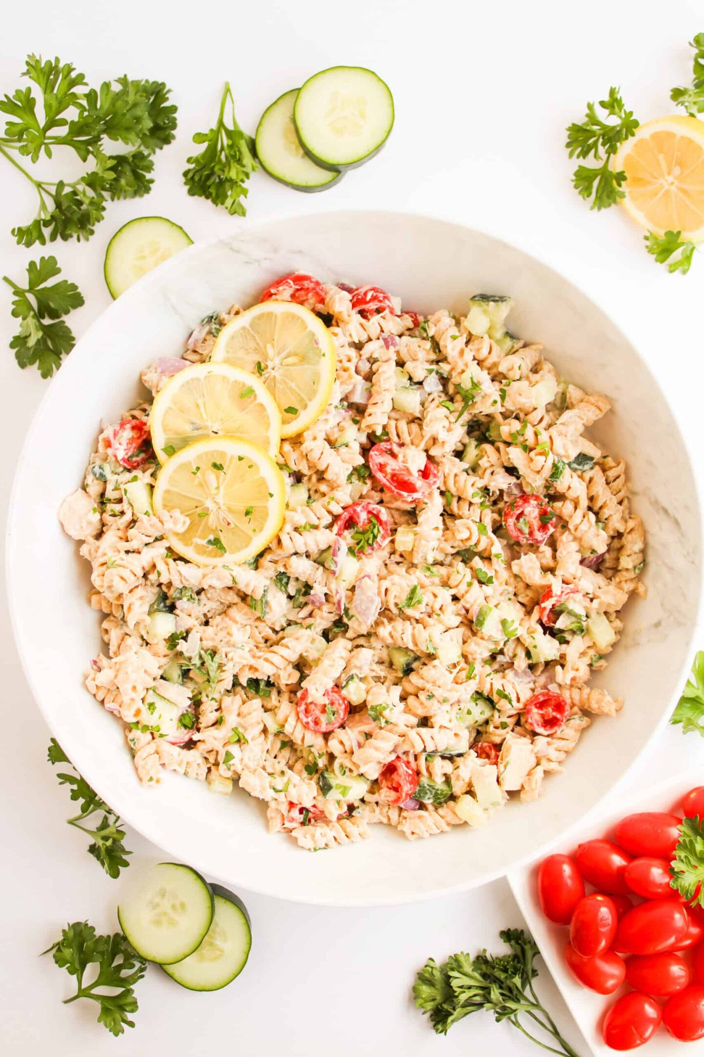 A white bowl of pasta salad with rotini, cherry tomatoes, cucumber, herbs, and lemon slices—perfect for side dishes at a graduation party—surrounded by fresh parsley, cucumber slices, lemon, and cherry tomatoes on a white surface.