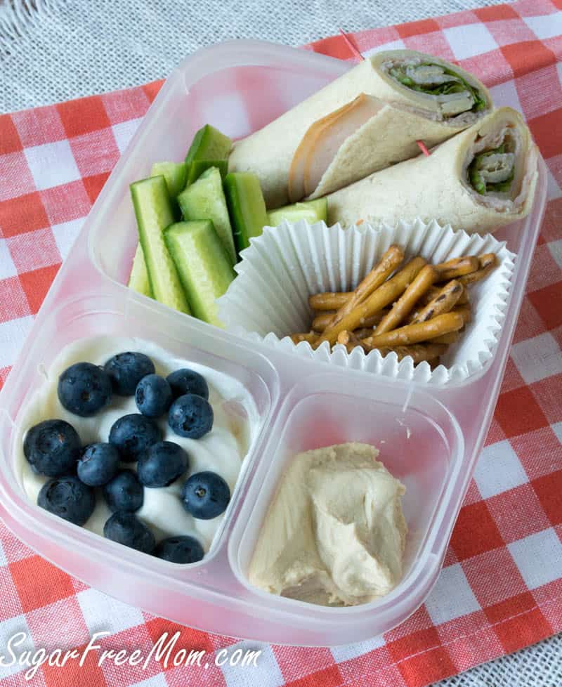 A kids lunch featuring turkey wrap halves, cucumber sticks, pretzel sticks, yogurt with blueberries, and hummus is served on a red and white checkered cloth—perfect for pool lunch ideas or easy pool snacks.