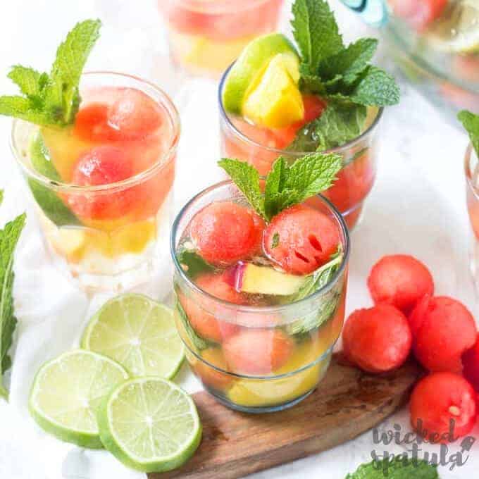 Three glasses filled with fruit balls, lime slices, and mint leaves on a board evoke the refreshing look of summer sangria, with extra limes and fruit balls scattered beside them.