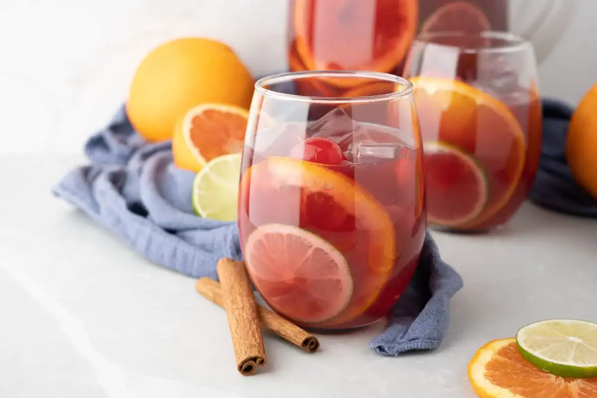 A glass of iced fruit drink with slices of orange, lime, and grapefruit, garnished with two cinnamon sticks—perfect inspiration for Fall Punch Recipes—surrounded by fresh citrus fruits and a blue cloth.