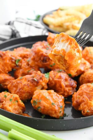 A fork holding a cut buffalo boneless wing over sauced chicken with celery and fries.