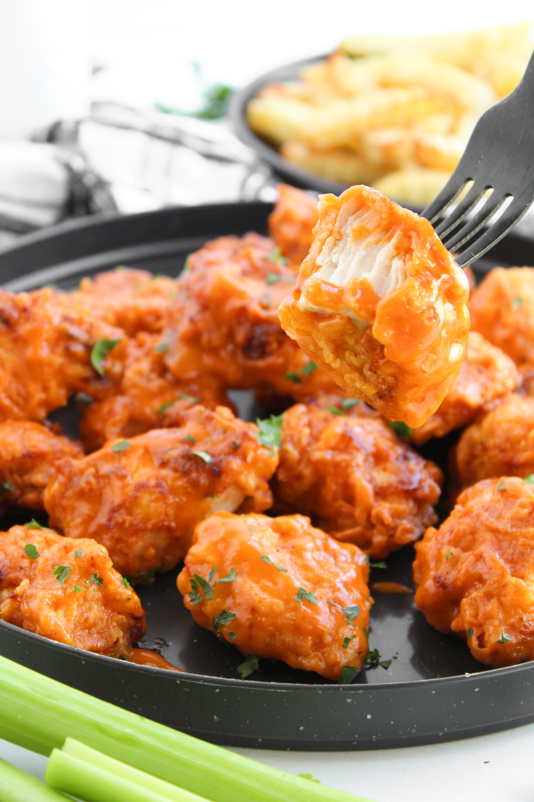 A fork holding a cut buffalo boneless wing over sauced chicken with celery and fries.