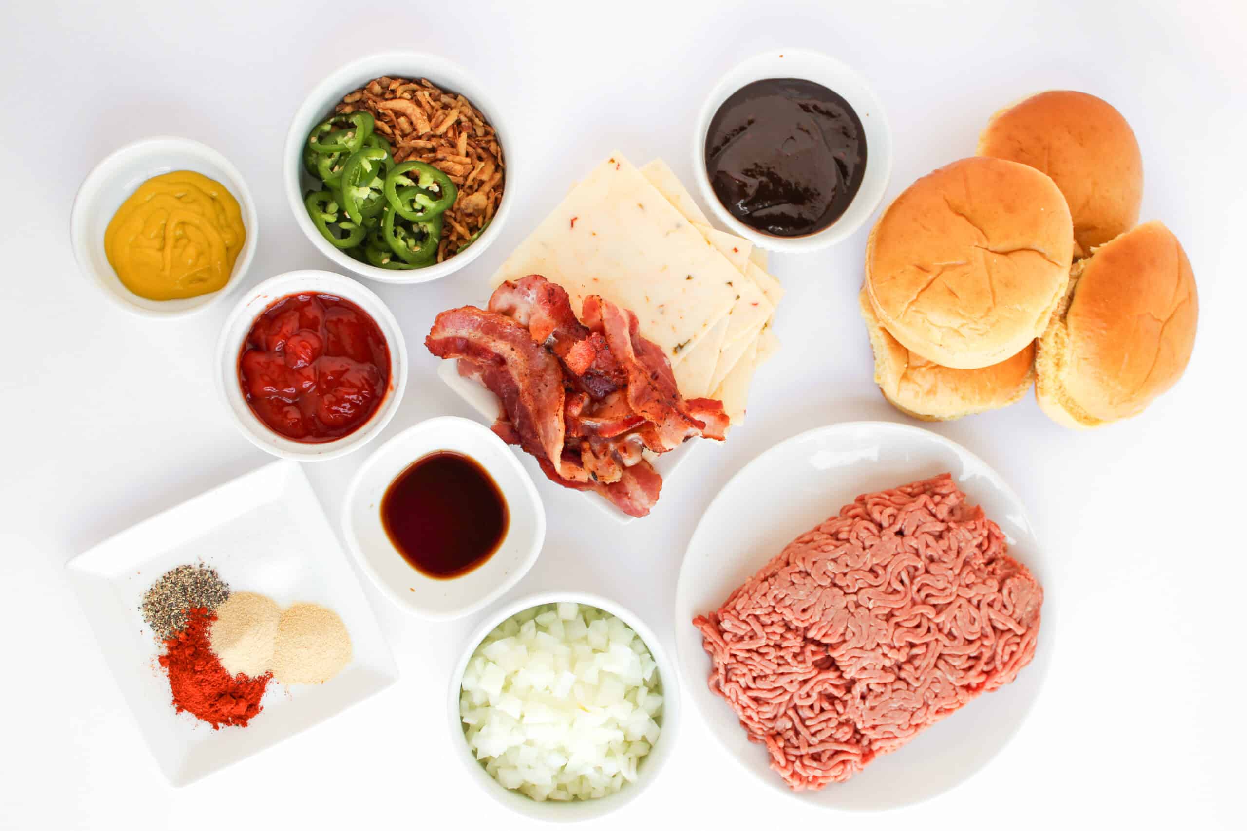 Ground beef, buns, bacon, cheese, sauces, seasonings, onion, jalapeño, and crispy onions arranged on a white surface.