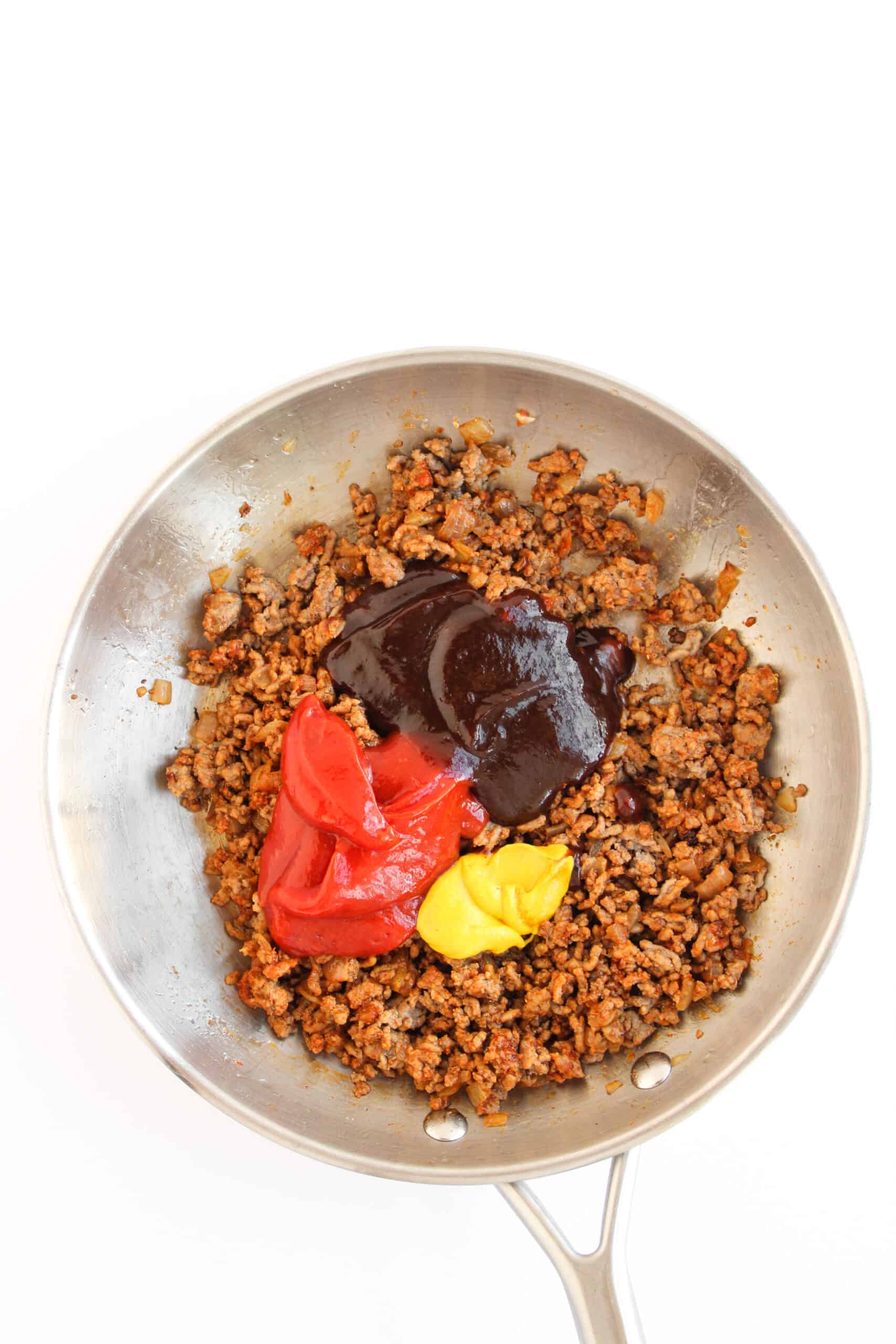 Barbecue sauce, ketchup, and mustard added to seasoned ground beef in a skillet.