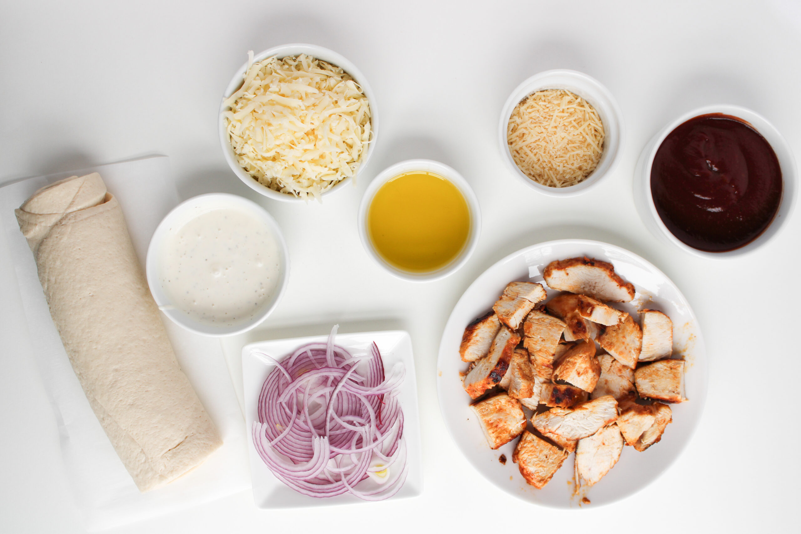 Pizza dough, cooked chicken, barbecue sauce, cheese, red onion, olive oil, and ranch arranged on a white surface.