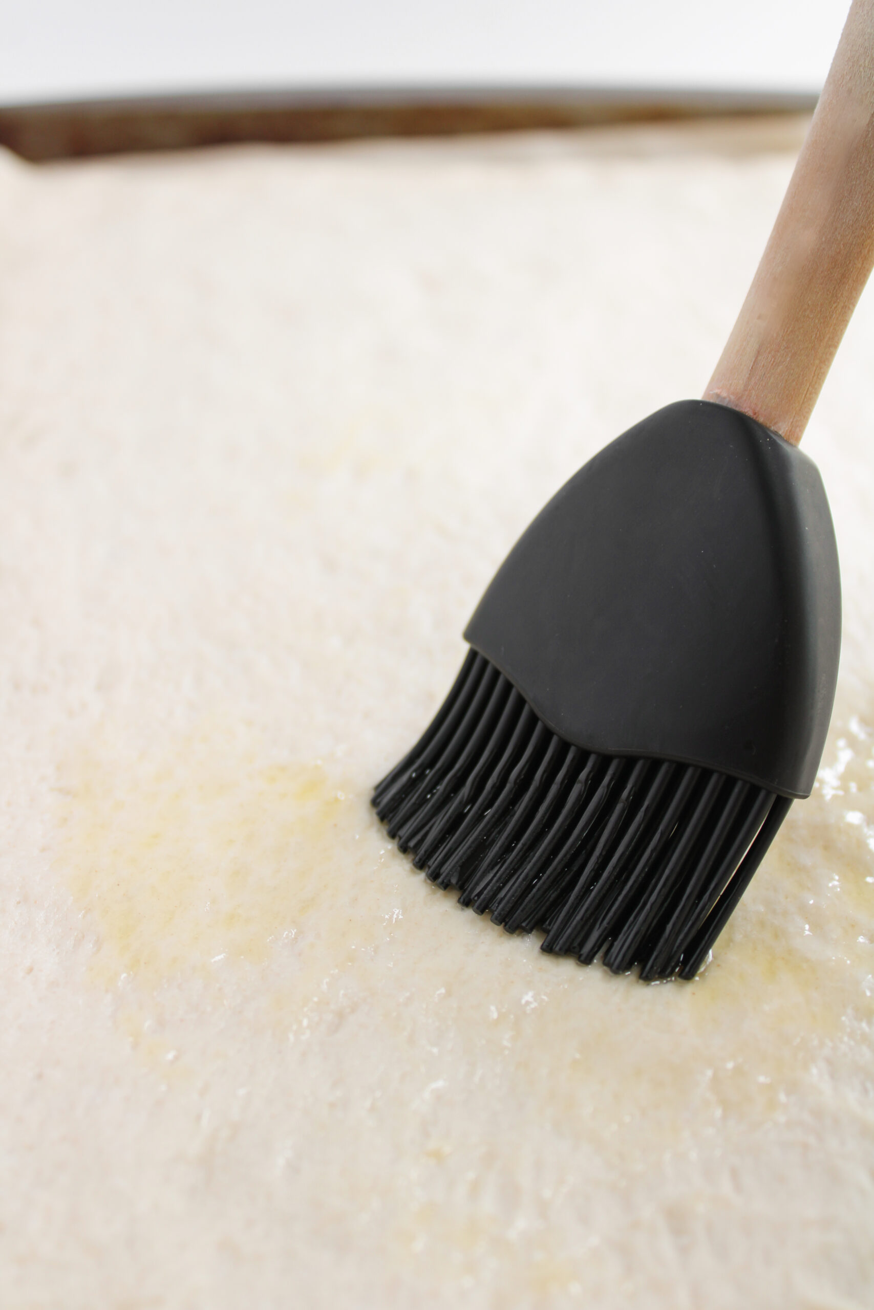 A pastry brush spreading olive oil over pizza dough in a sheet pan.