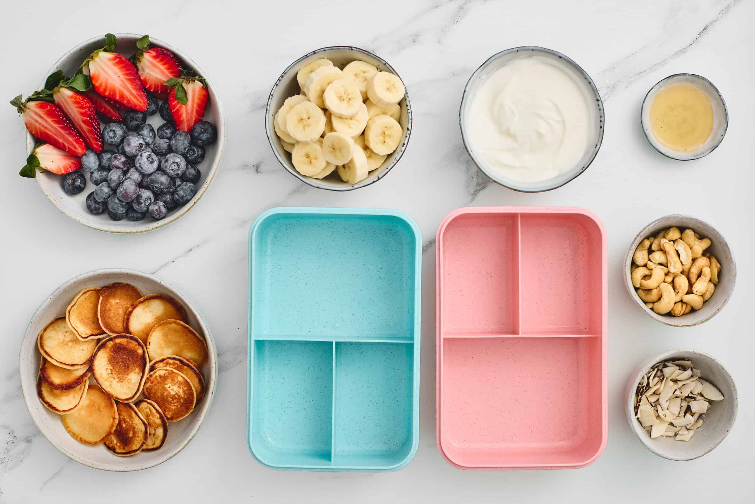 Mini pancakes, berries, banana, yogurt, honey, cashews, and coconut beside bento boxes.
