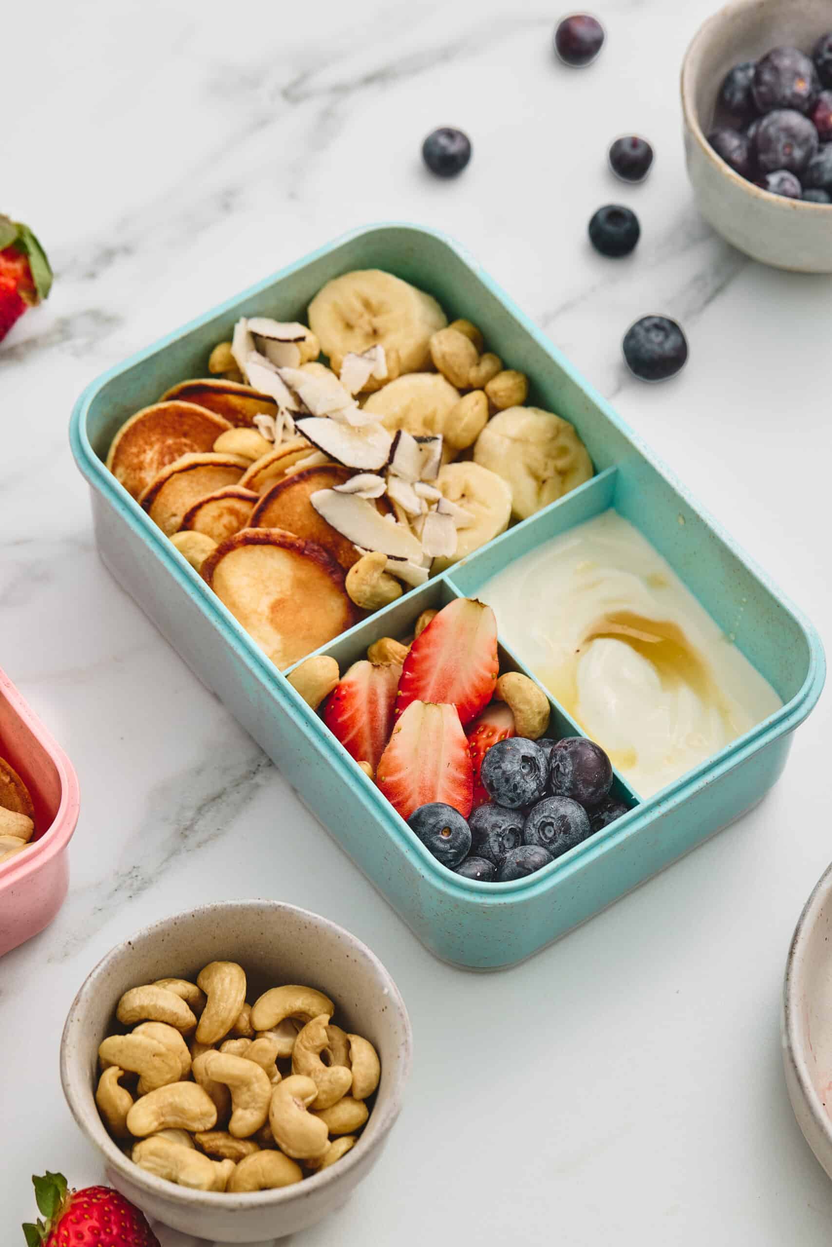 Close-up of a breakfast lunchbox filled with mini pancakes, fruit, yogurt, cashews, and coconut.