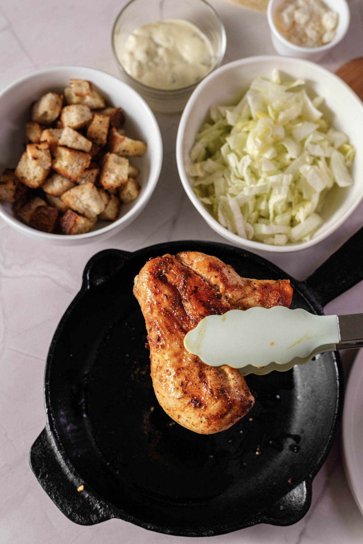 Grilled chicken breast held by tongs over a skillet, with bowls of croutons, chopped lettuce, and dressing on a countertop—perfect ingredients for assembling delicious Chicken Caesar Wraps.