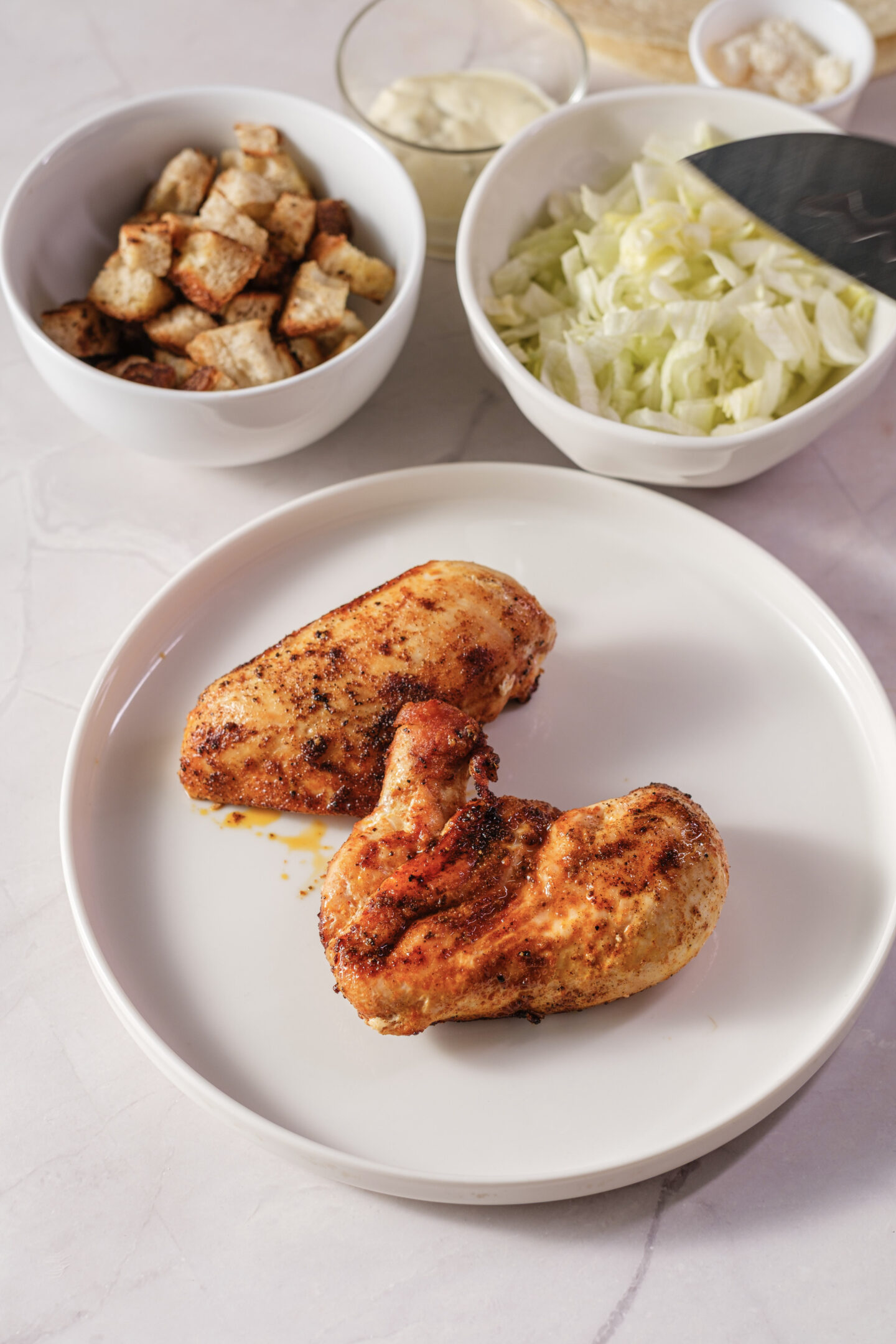 Two seasoned, cooked chicken breasts on a white plate with bowls of croutons, chopped lettuce, and condiments in the background—perfect ingredients for assembling flavorful Chicken Caesar Wraps.