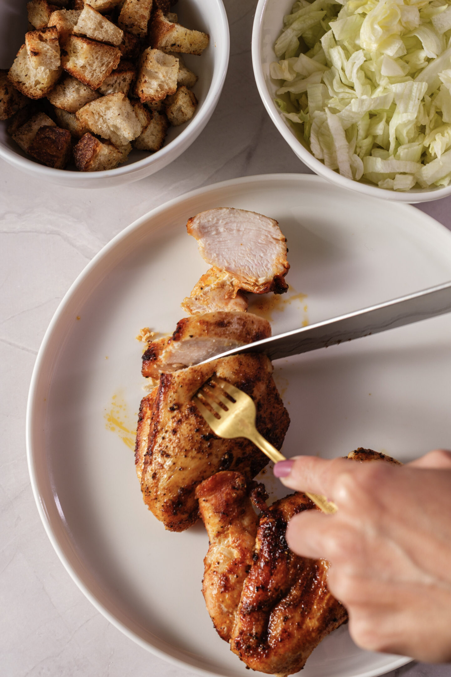 A person slices cooked chicken breast on a plate, with bowls of croutons and shredded lettuce nearby on a countertop—perfect ingredients for delicious Chicken Caesar Wraps.
