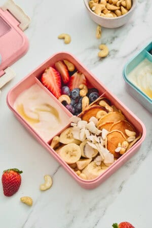 Mini pancake and fruit breakfast lunchbox with yogurt, honey, cashews, and coconut.