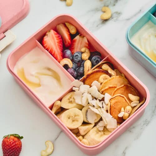 Mini pancake and fruit breakfast lunchbox with yogurt, honey, cashews, and coconut.