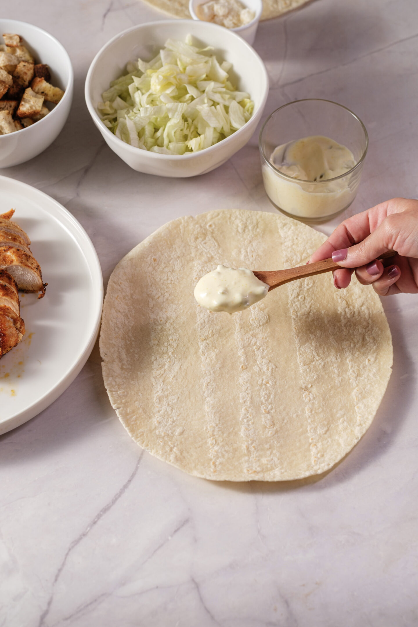 A hand spreads creamy dressing onto a tortilla wrap, preparing the base for delicious Chicken Caesar Wraps, with bowls of lettuce, croutons, and sliced chicken arranged nearby on a light countertop.