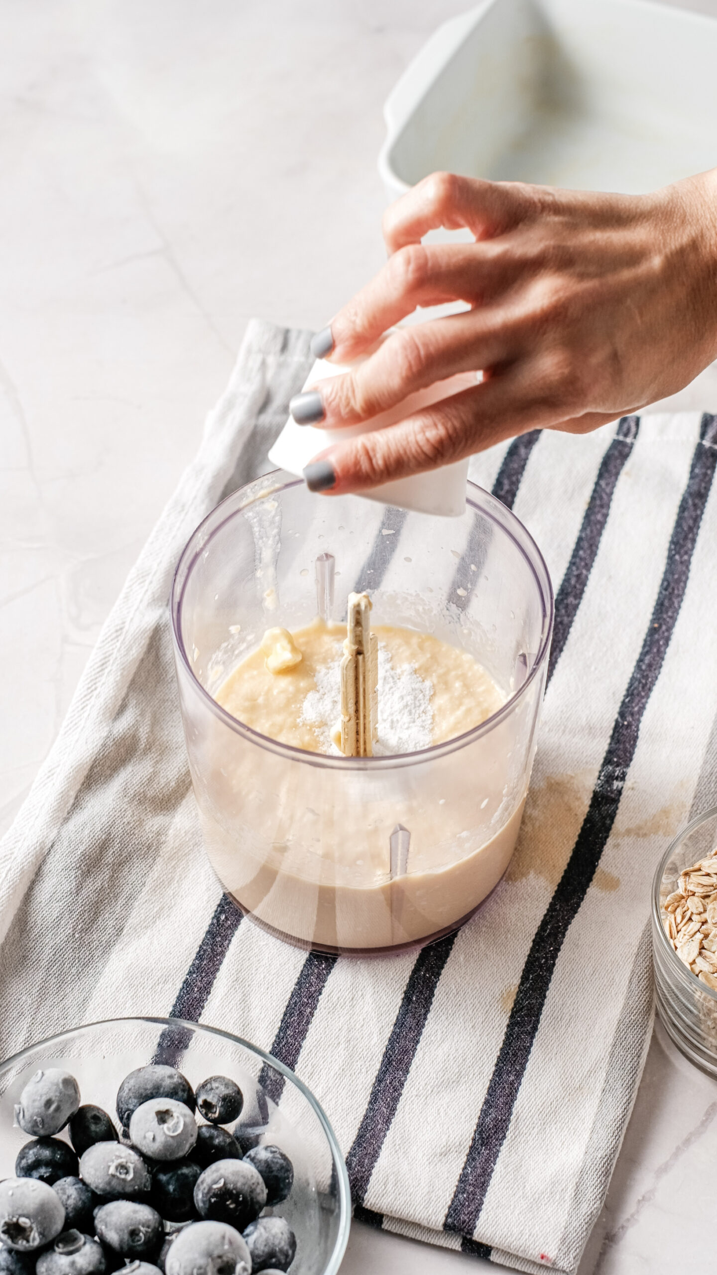 A hand adds a white ingredient, possibly cottage cheese, to a food processor containing a beige mixture, with blueberries and oats in bowls nearby on a striped kitchen towel—perfect for preparing a wholesome blueberry breakfast bake.