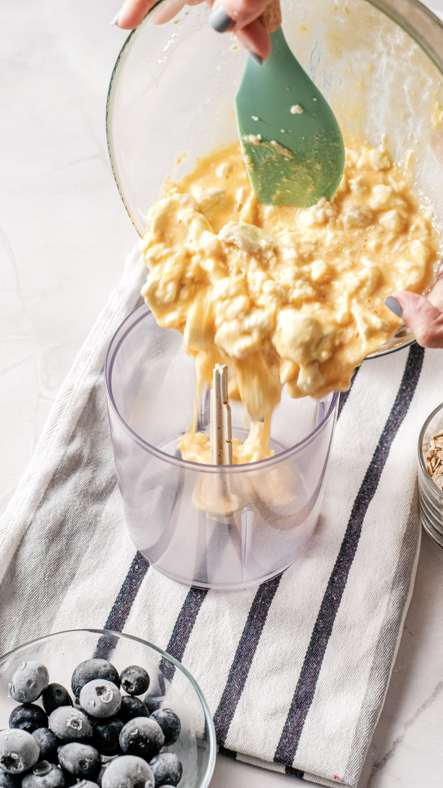Hands pour a chunky yellow cottage cheese batter from a bowl into a blender jar on a striped towel, with a bowl of blueberries and a small dish of oats nearby—perfect for preparing a delicious blueberry breakfast bake.