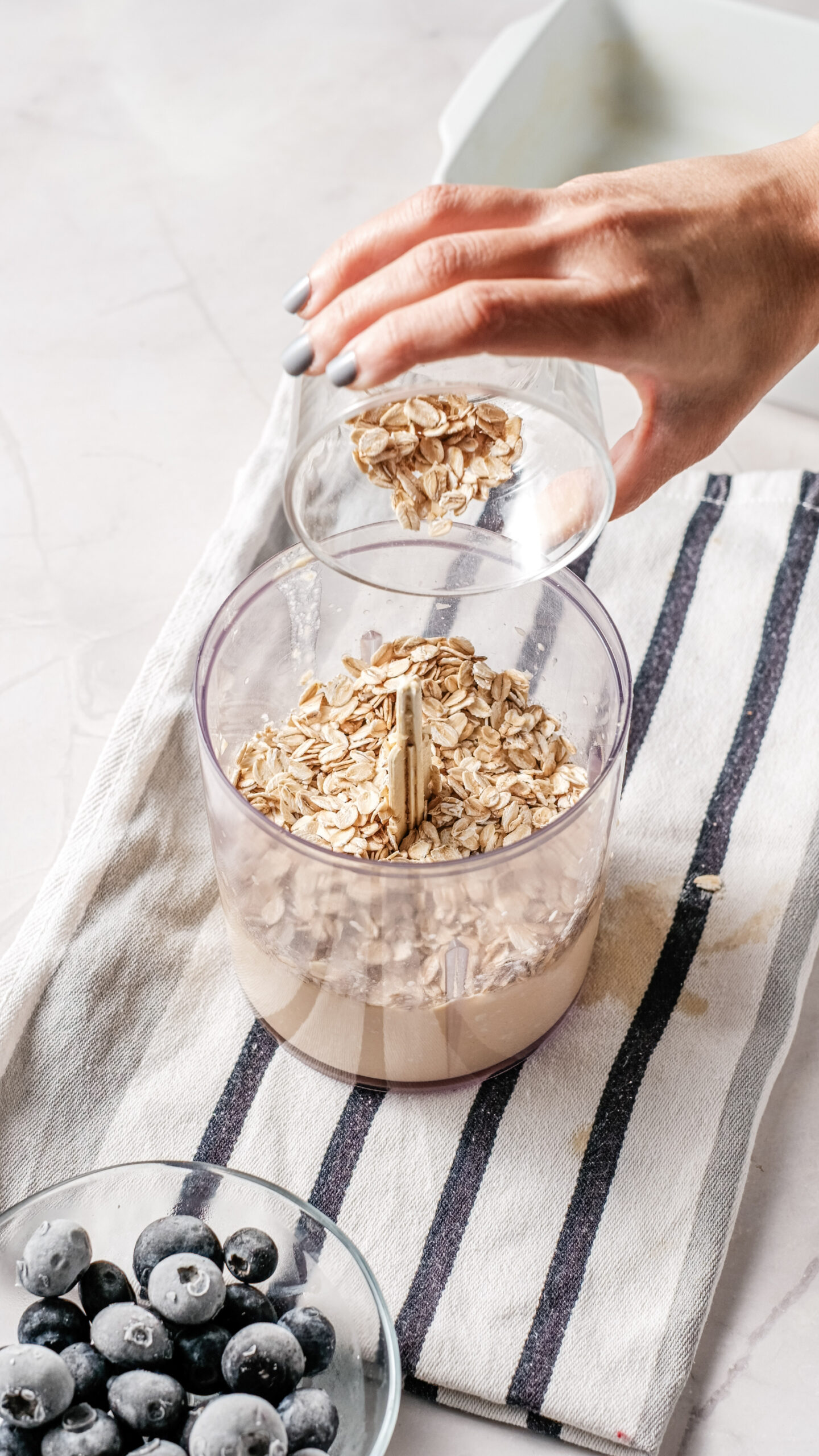 A hand pours oats from a small glass bowl into a food processor containing oats and liquid, preparing the base for a wholesome Breakfast Bake, with fresh blueberries and a hint of creamy cottage cheese nearby on a striped towel.