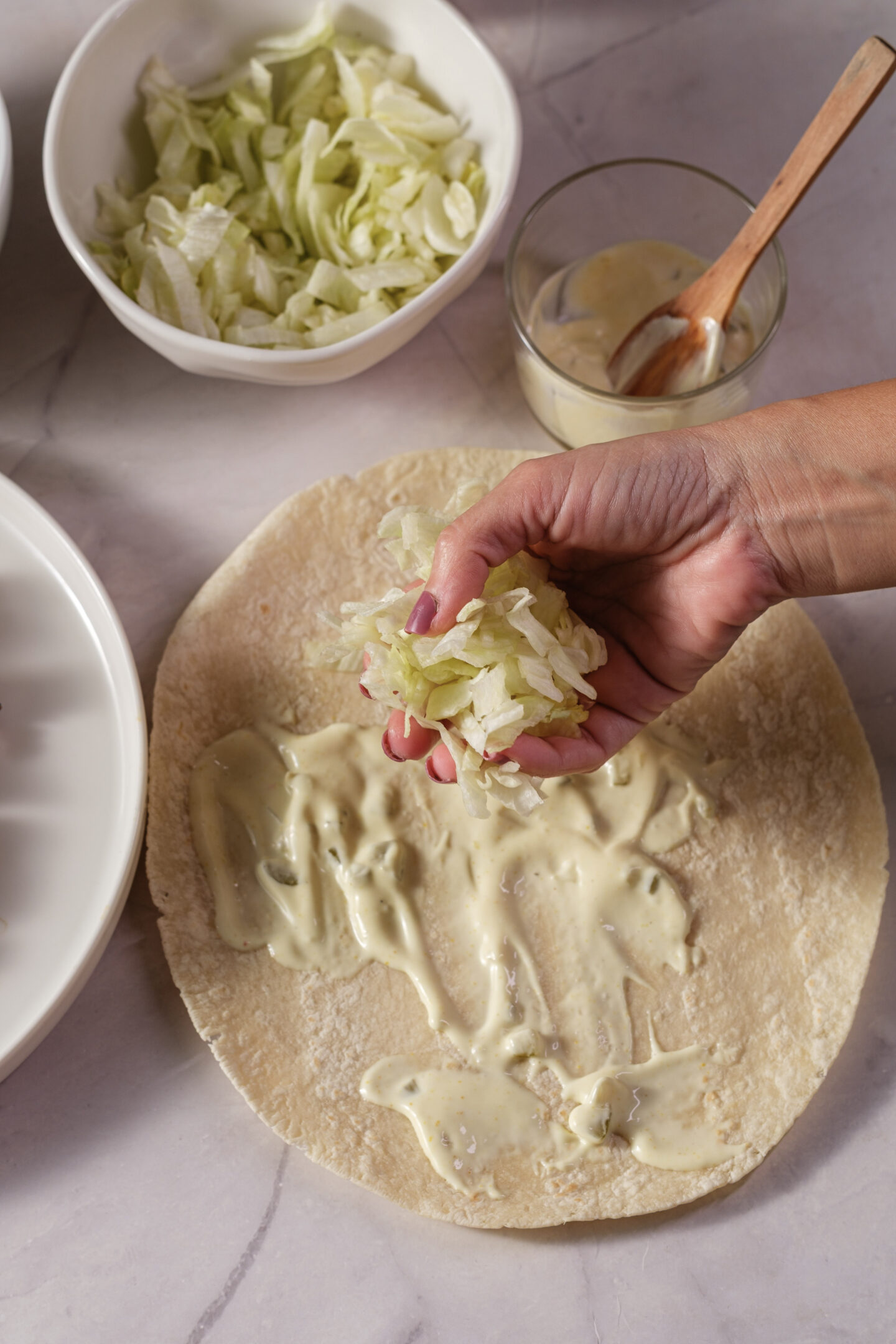 A hand adds shredded lettuce to a tortilla spread with sauce, preparing the base for delicious Chicken Caesar Wraps, with bowls of lettuce and sauce visible nearby on a countertop.