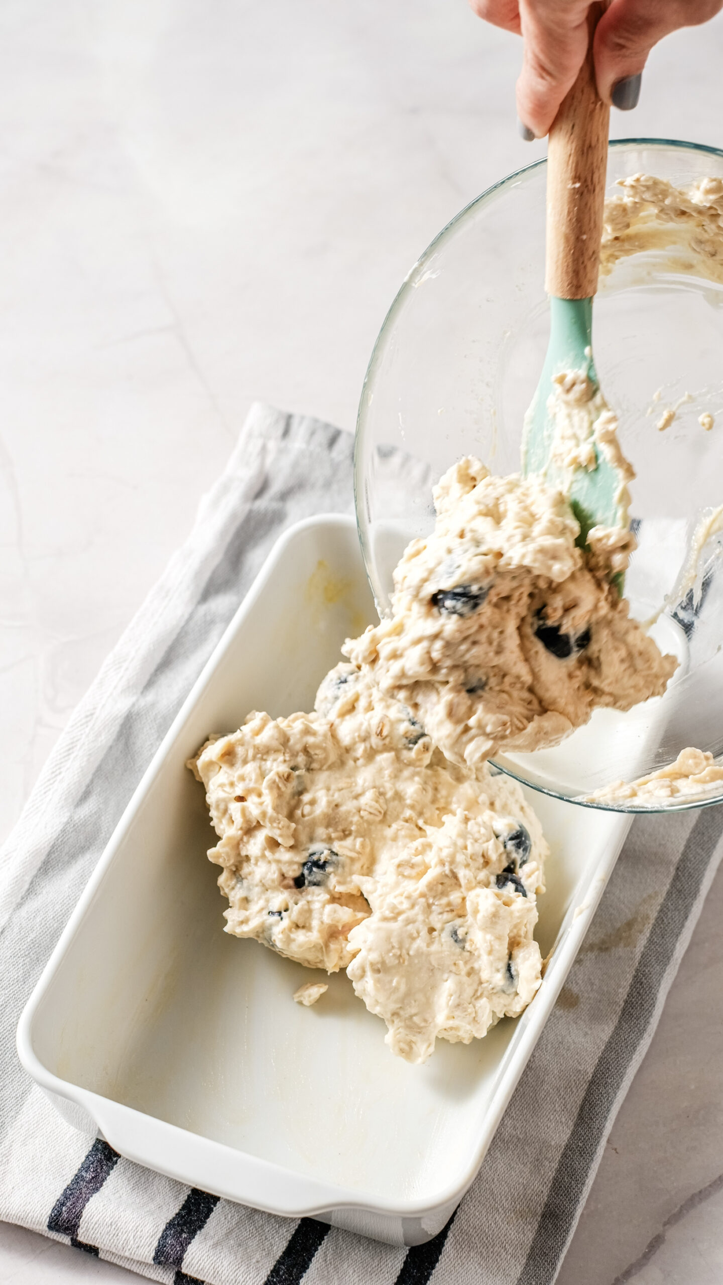 A hand uses a spatula to transfer a chunky, light-colored cottage cheese batter with dark blueberry pieces from a glass bowl into a rectangular baking dish on a striped cloth, preparing it for a delicious breakfast bake.