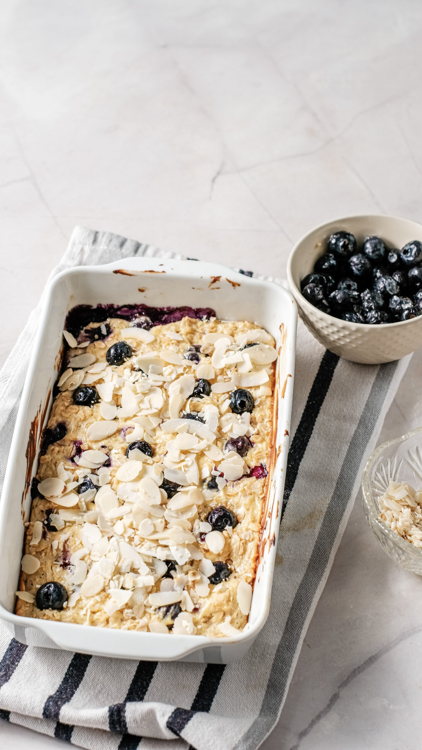 Baked oatmeal breakfast bake topped with juicy blueberries and sliced almonds in a white dish, with bowls of fresh blueberries and almonds beside it on a striped cloth. This wholesome treat is perfect for pairing with creamy cottage cheese to start your day right.