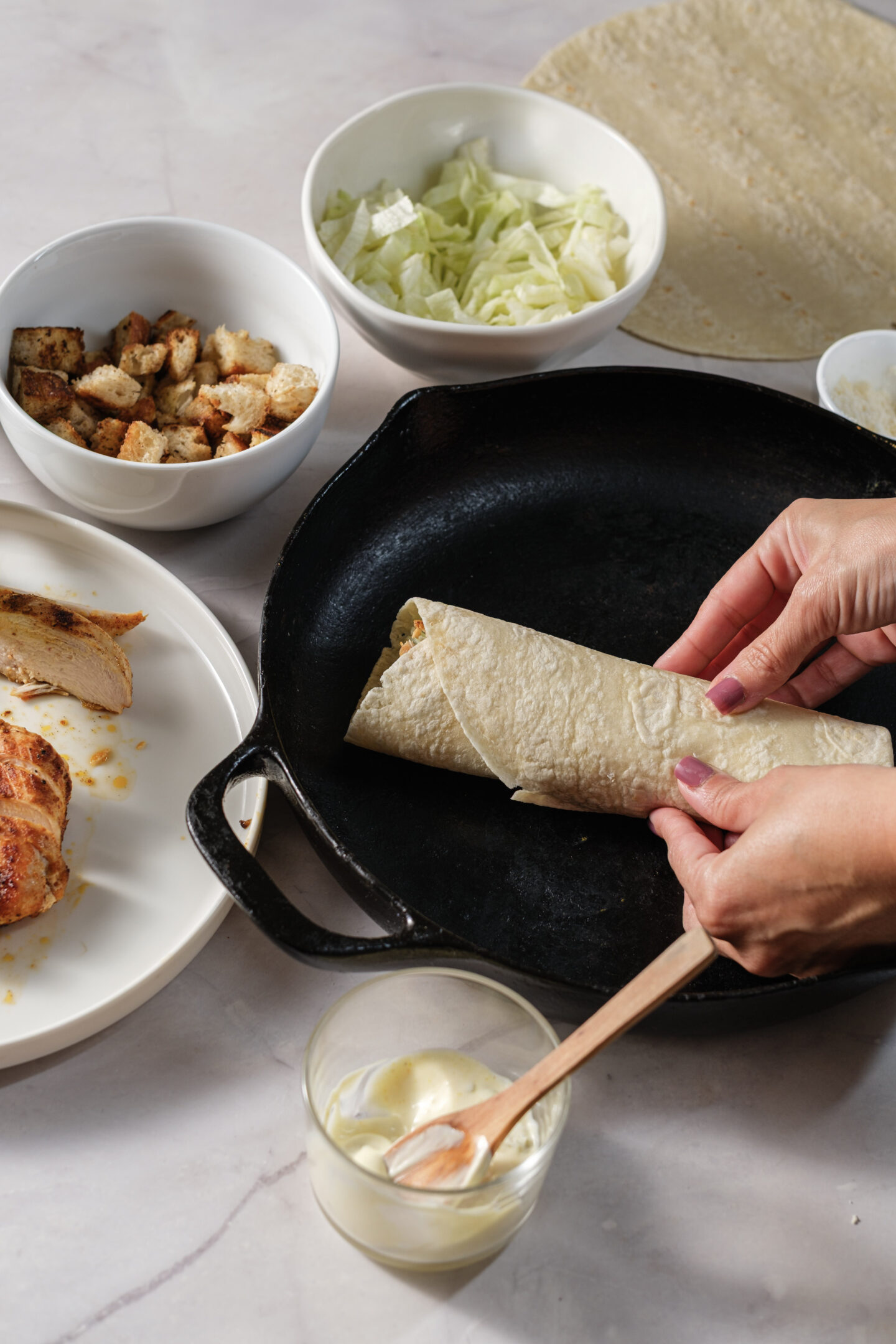 Hands rolling a Chicken Caesar Wrap with tender chicken, crisp lettuce, and creamy sauce next to a cast iron pan, with bowls of ingredients on a light countertop.