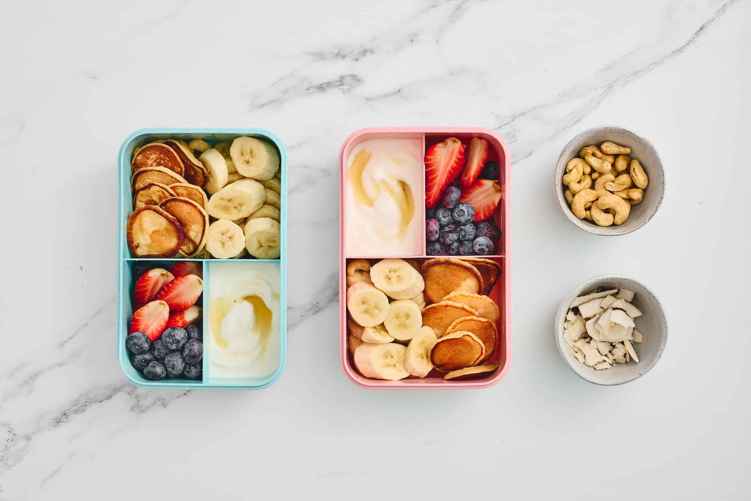 Two assembled lunchboxes with mini pancakes, fruit, yogurt, cashews, and coconut.