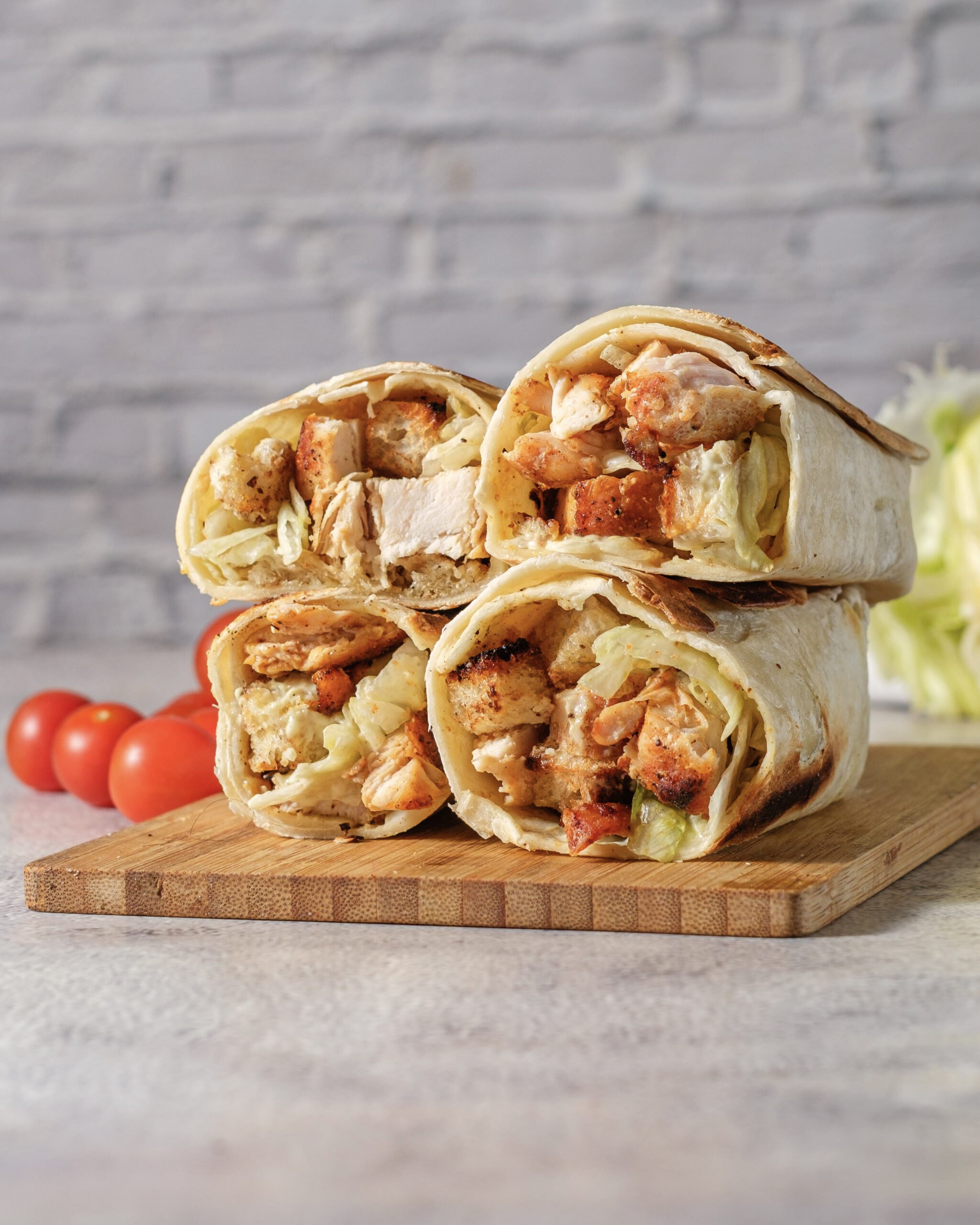 Four toasted chicken Caesar wrap halves stacked on a cutting board with tomatoes and romaine.