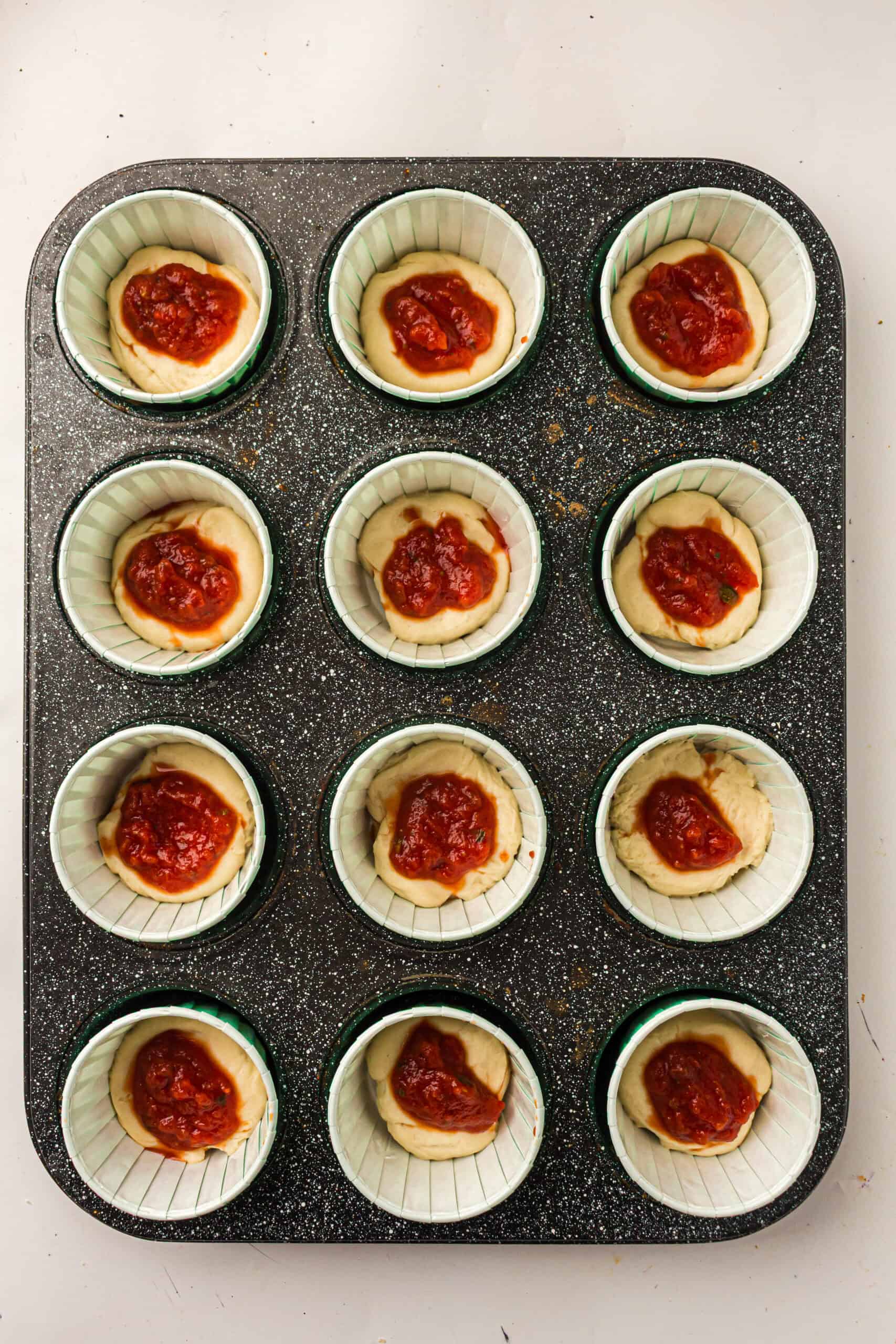 Pizza dough portions topped with thick pizza sauce in a muffin tin.