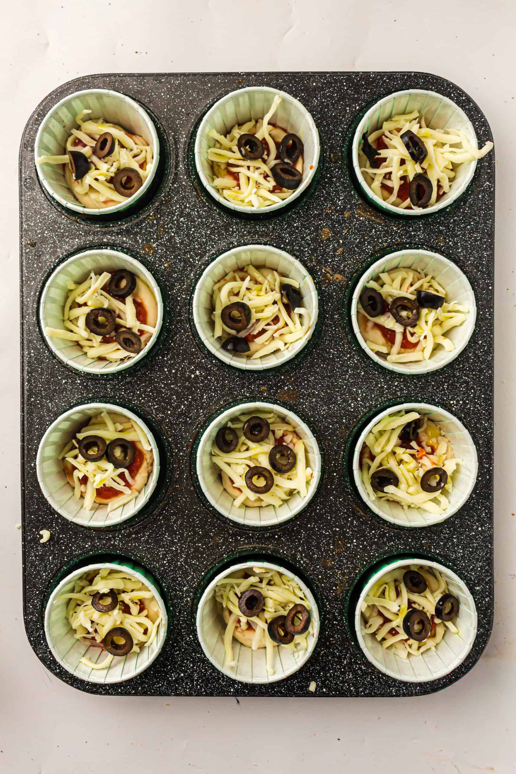 Unbaked pizza muffins topped with mozzarella and sliced black olives.