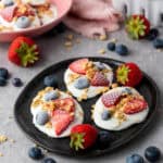 Frozen vanilla yogurt bites with strawberries, blueberries, and granola on a black plate.
