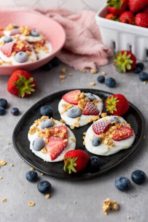 Frozen vanilla yogurt bites with strawberries, blueberries, and granola on a black plate.