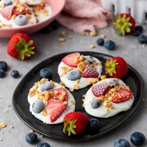 Frozen vanilla yogurt bites with strawberries, blueberries, and granola on a black plate.