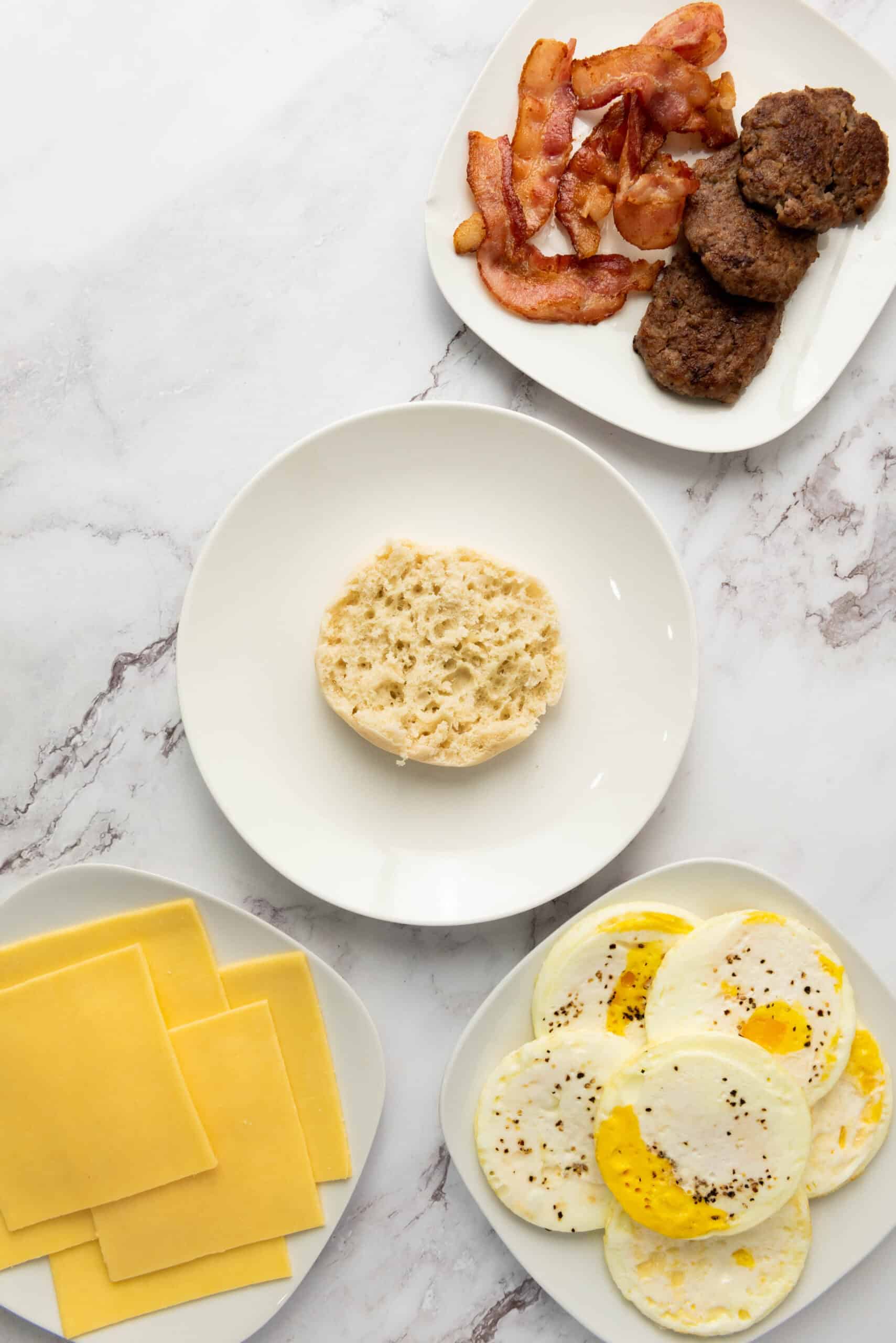 Cooked eggs, bacon, sausage, cheese, and an English muffin set out for sandwich assembly.