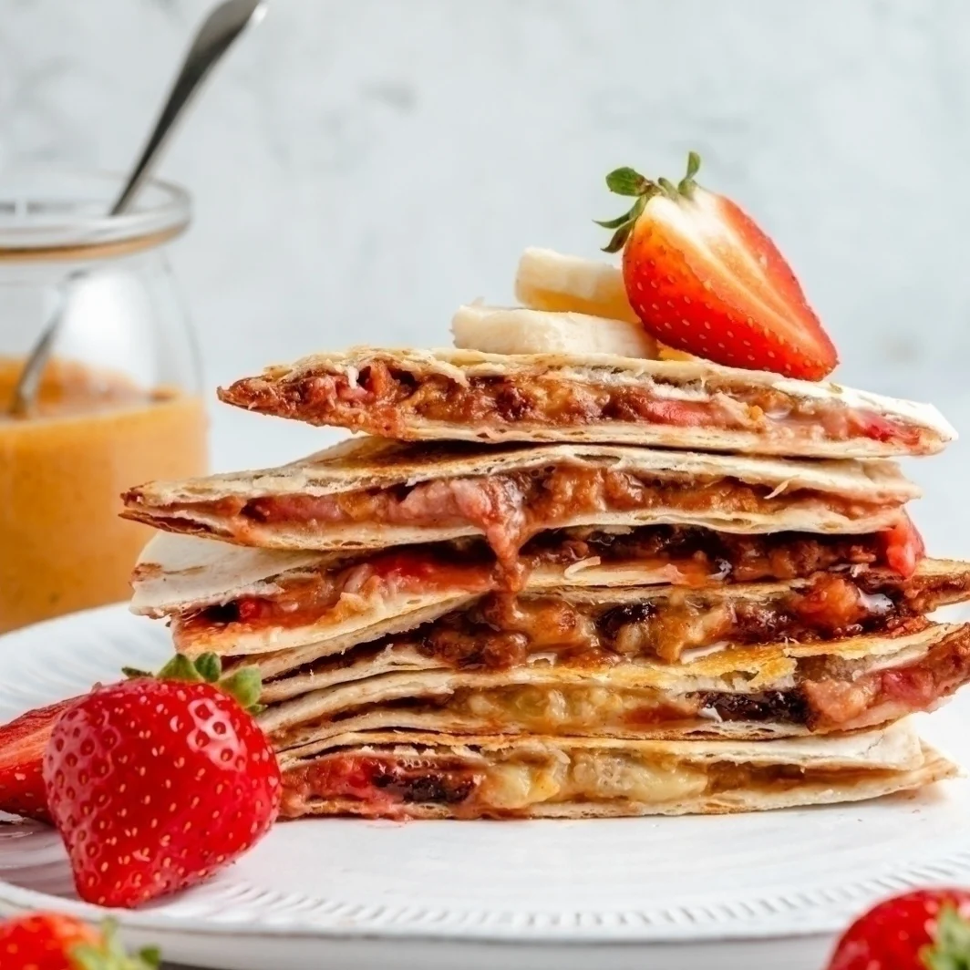 Stack of quesadillas filled with peanut butter, banana, and strawberries on a white plate, garnished with fresh strawberries and banana slices. Perfect as after-school snacks or fun snacks for kids, this colorful treat is sure to be a hit. A jar of peanut butter is in the background.