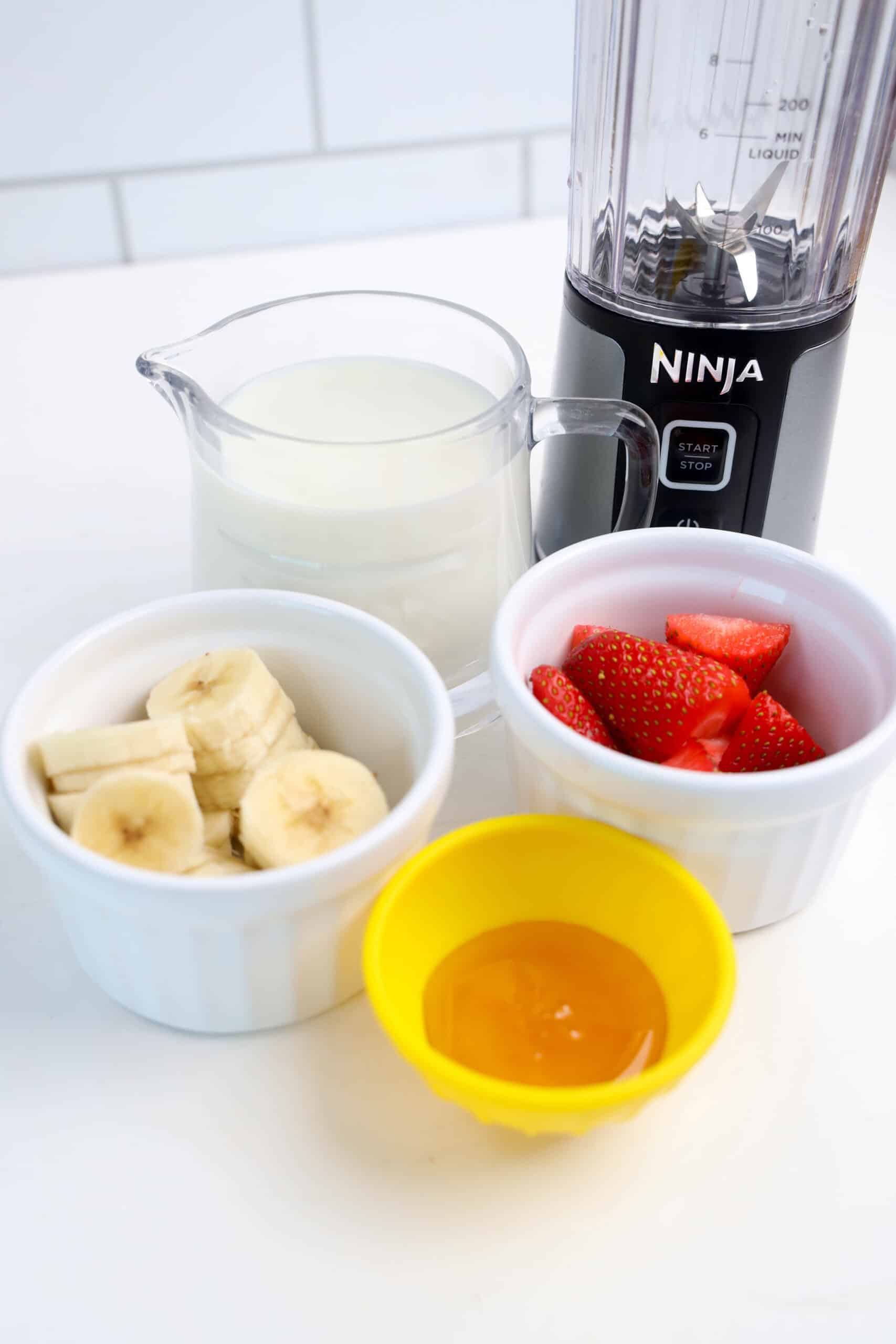 Milk, sliced banana, strawberries, and honey beside a personal blender.