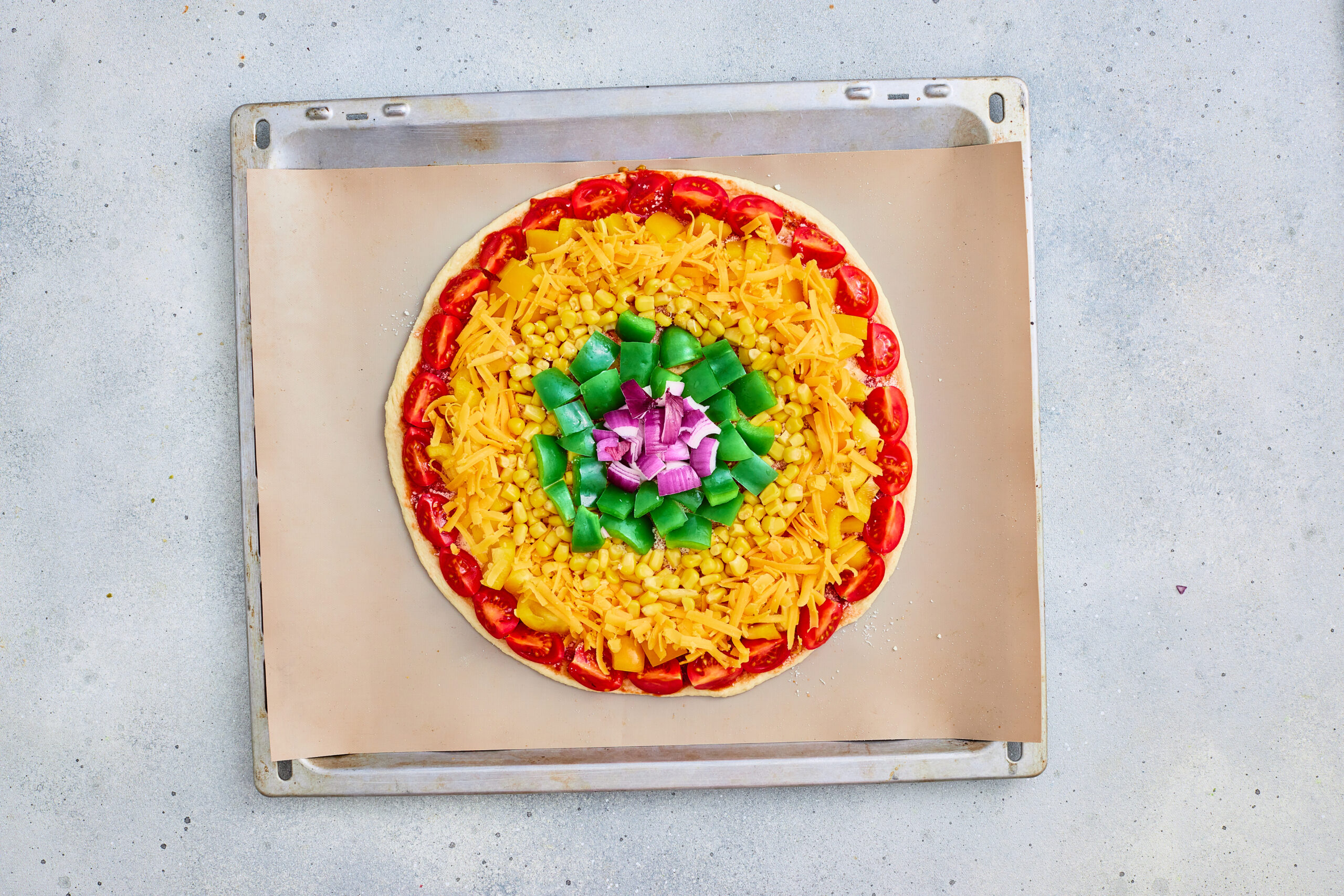 Completed unbaked rainbow pizza with tomato, yellow pepper, cheddar, corn, green pepper, and red onion rings.