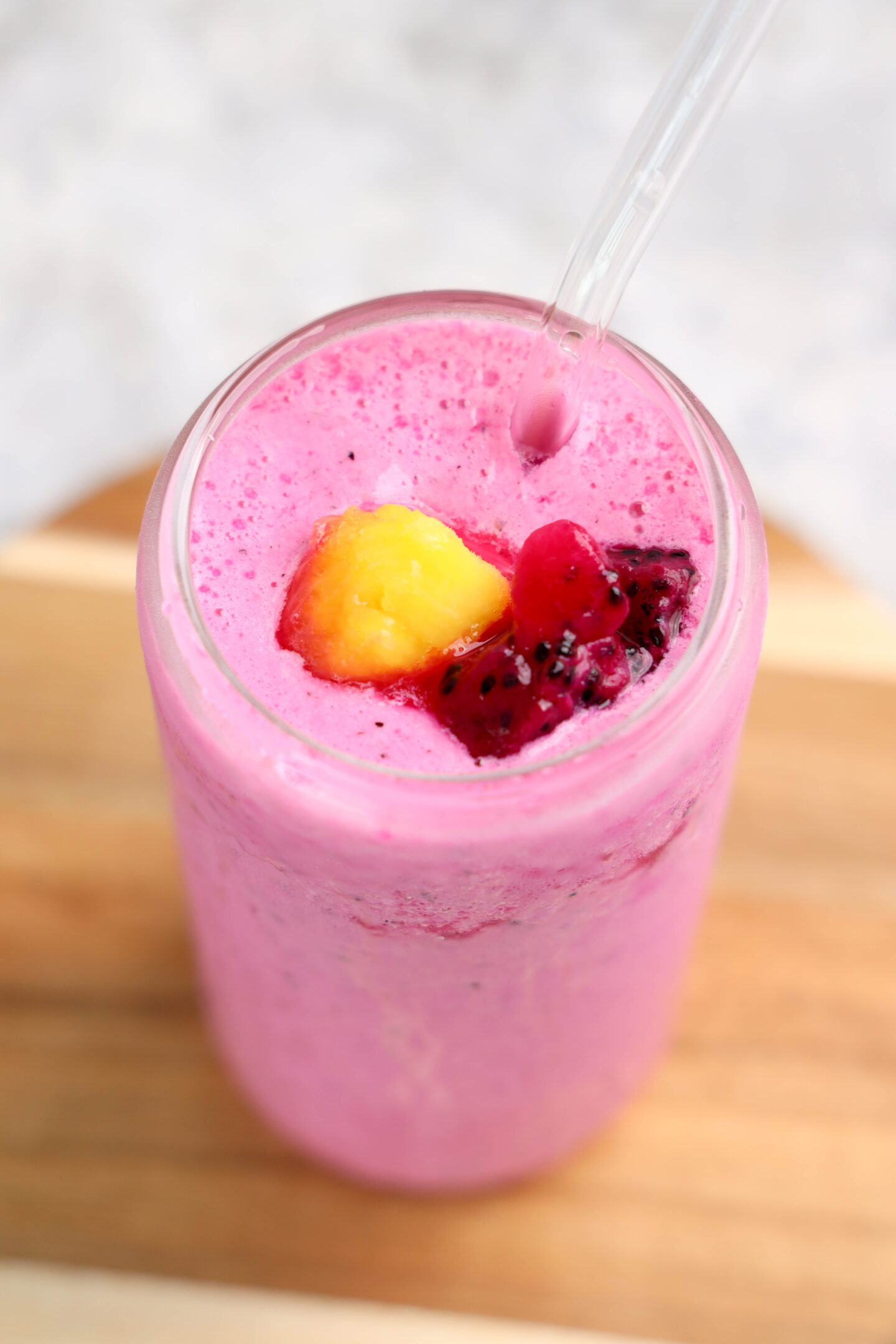 A glass of bright pink dragonfruit smoothie with a clear straw, topped with diced yellow and red fruit pieces, placed on a wooden surface.