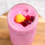 A glass of bright pink dragonfruit smoothie with a clear straw, topped with diced yellow and red fruit pieces, placed on a wooden surface.