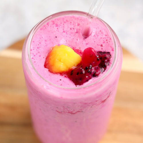 A glass of bright pink dragonfruit smoothie with a clear straw, topped with diced yellow and red fruit pieces, placed on a wooden surface.