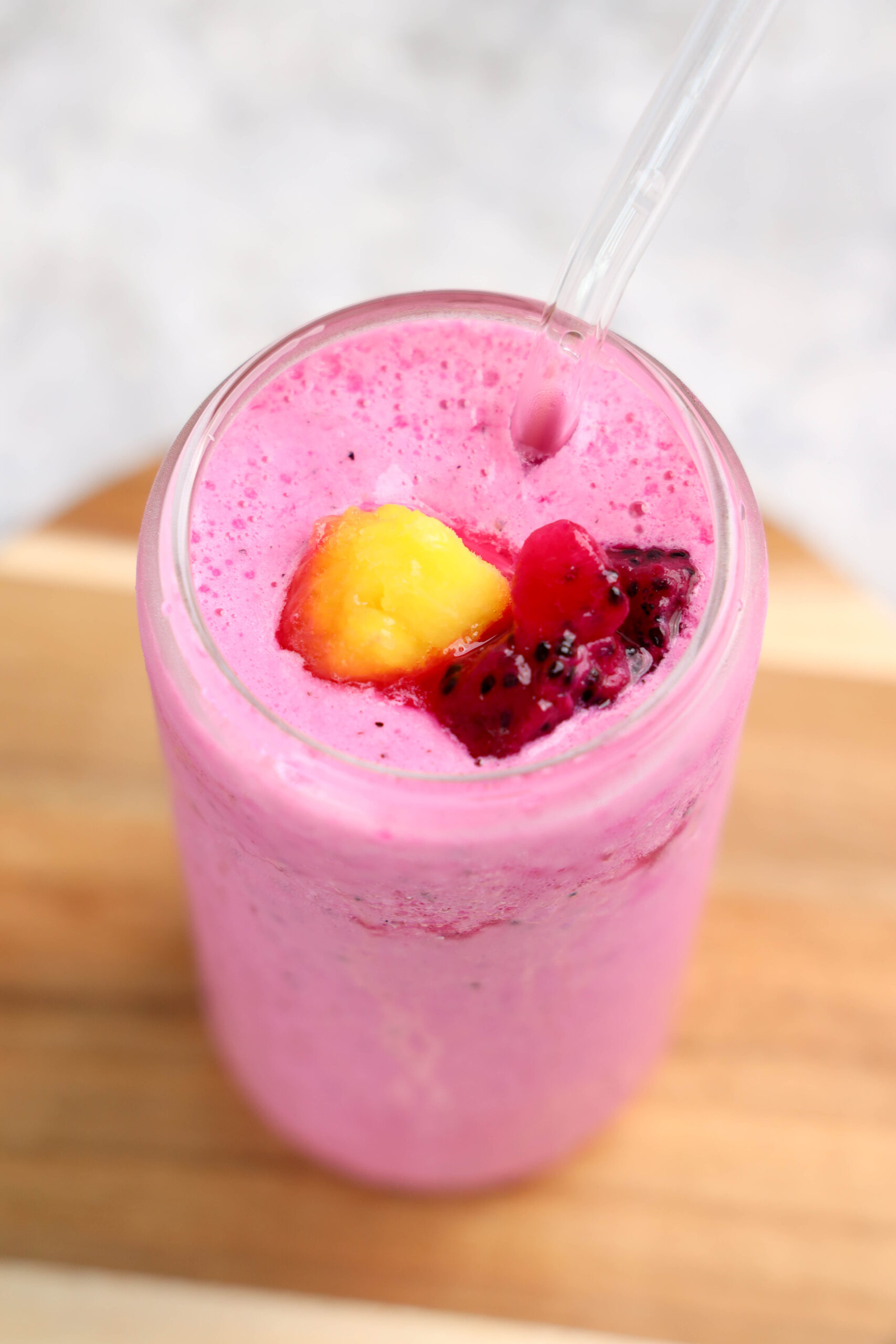 A glass of bright pink dragonfruit smoothie with a clear straw, topped with diced yellow and red fruit pieces, placed on a wooden surface.