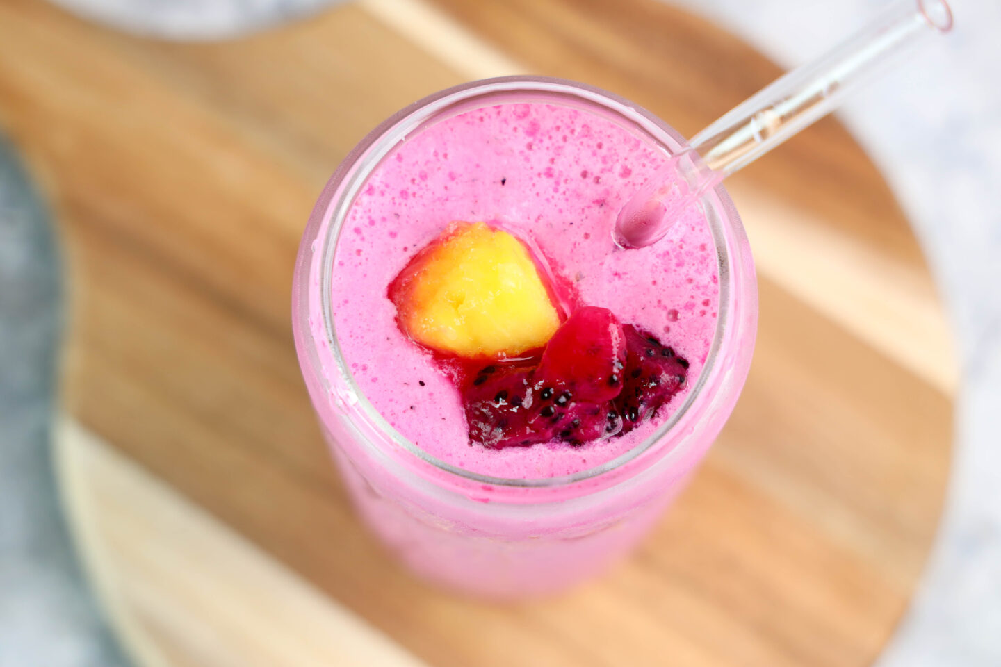 A vibrant Dragonfruit Smoothie in a glass, topped with pieces of yellow mango and red dragon fruit, with a clear straw, sits invitingly on a wooden surface.