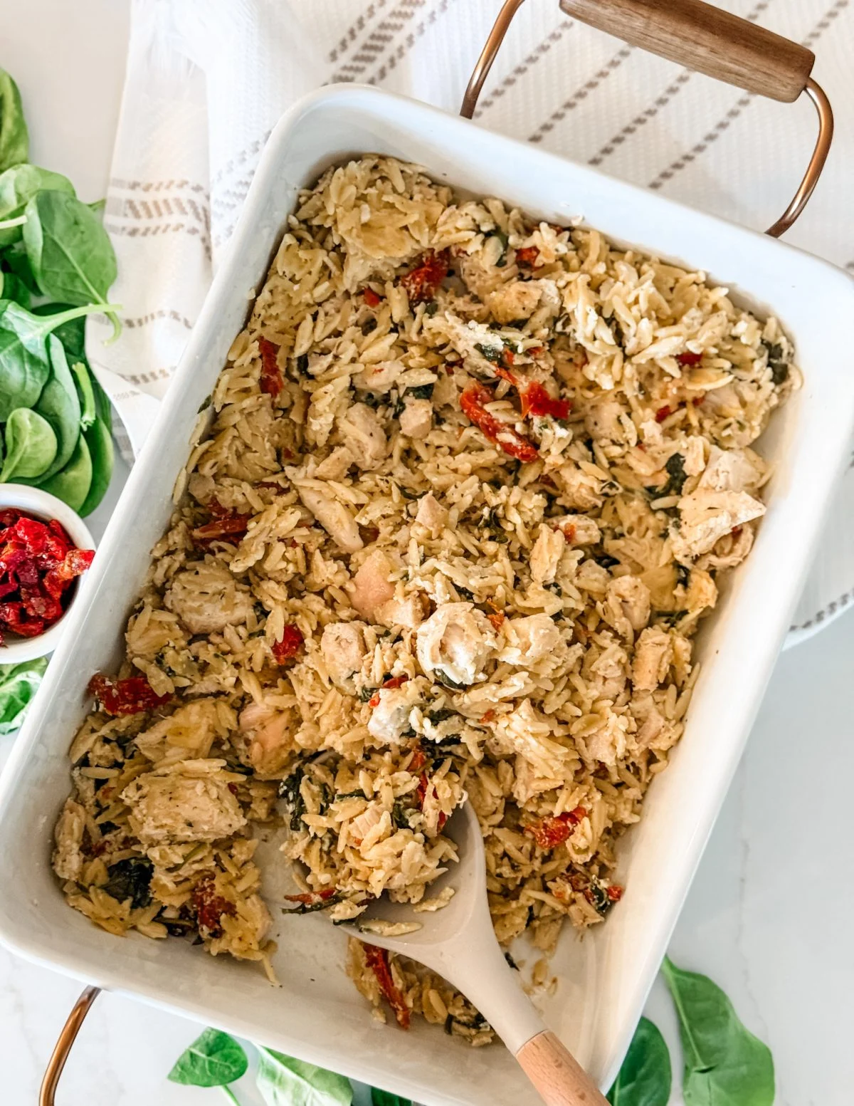 Baked chicken and orzo casserole with sun-dried tomatoes and spinach in a white dish, partially served with a spoon. This comforting meal is perfect for fans of dump and bake casseroles—simply add your ingredients to one pan, bake, and enjoy! Fresh spinach and a bowl of sun-dried tomatoes are on the side.