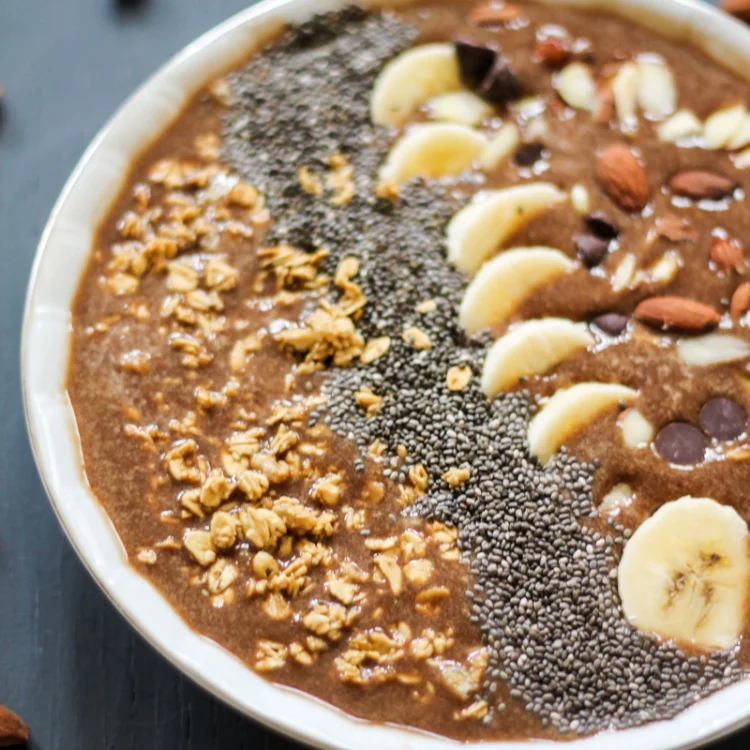 A high-protein smoothie bowl topped with banana slices, chia seeds, granola, almonds, and chocolate chips, arranged in rows on a white plate—perfect inspiration for your favorite smoothie recipes.