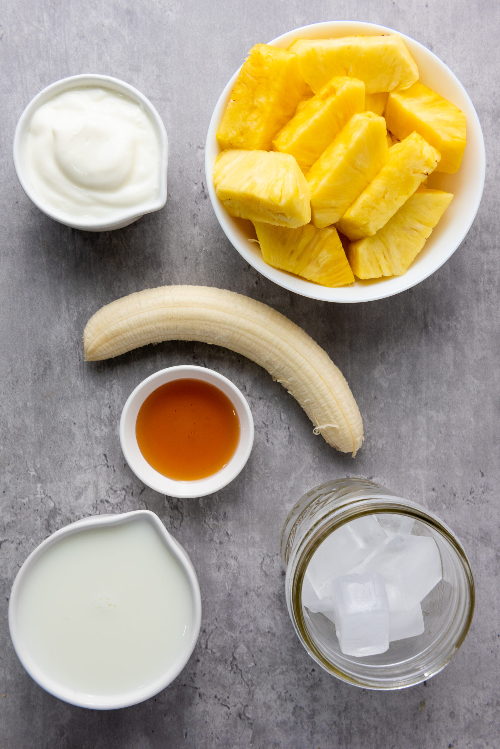 Pineapple, banana, Greek yogurt, milk, honey, and ice arranged on a gray surface.